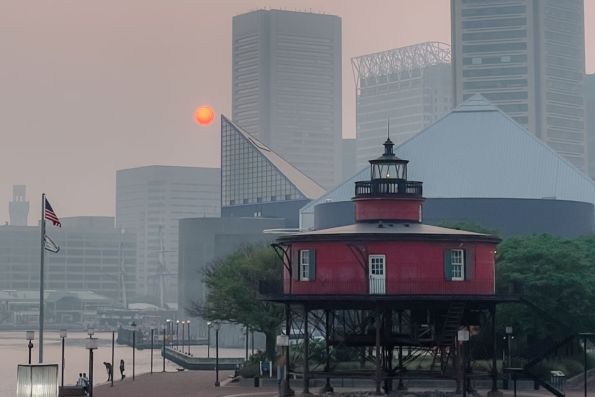 Smoky skies and an orange sun backdrop skyscrapers near a harbor.