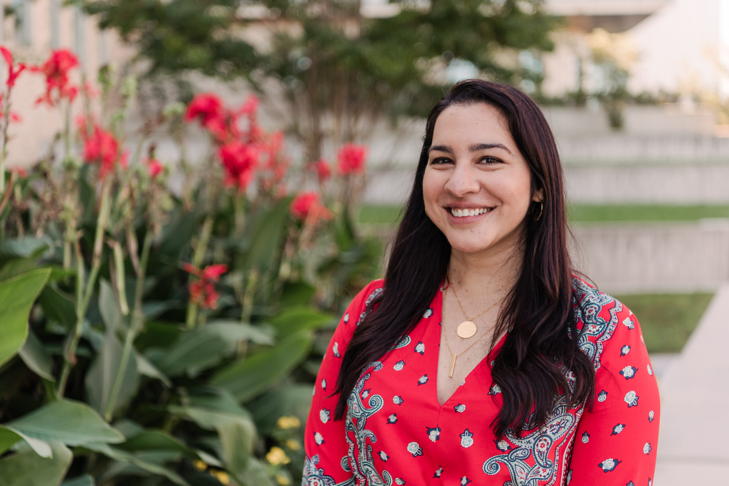 Noor Zaidi wearing a coral red blouse stands outside in front of a bed of tall flowers.
