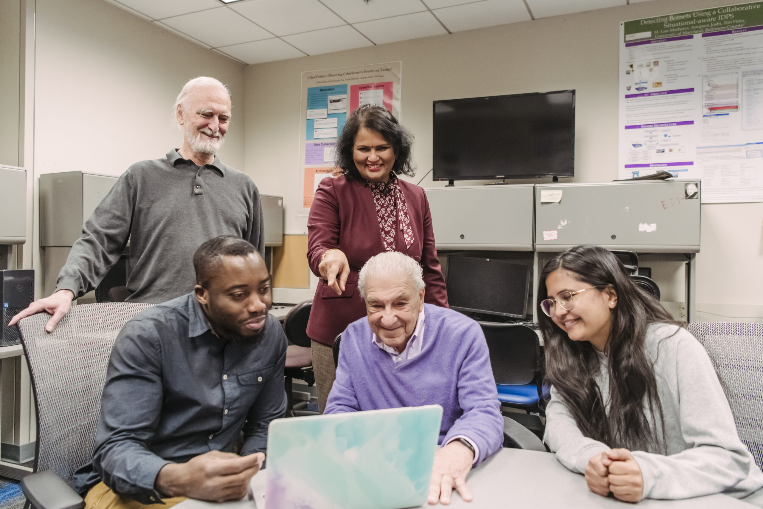 A group of people sitting and standing around a laptop in a classroom. One woman s pointing to the laptop.