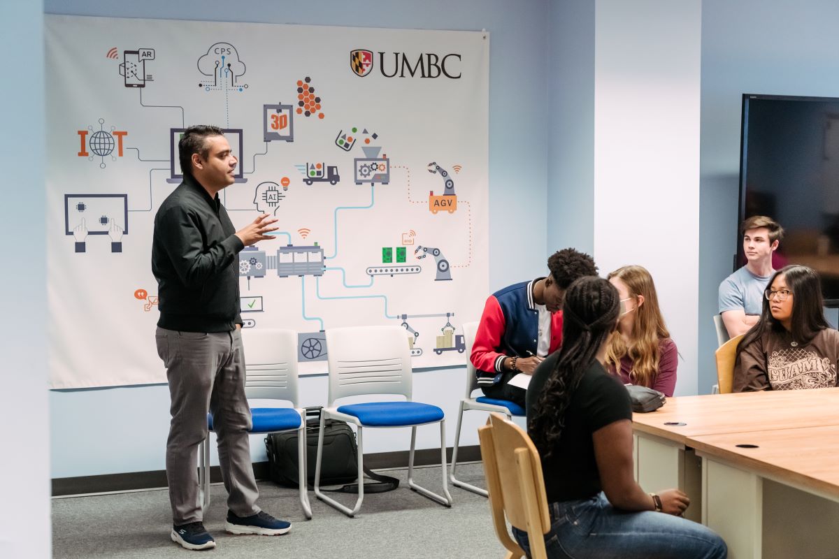 Man stands in front of poster with symbols of robots, trucks, computers and talks to group of seated students.