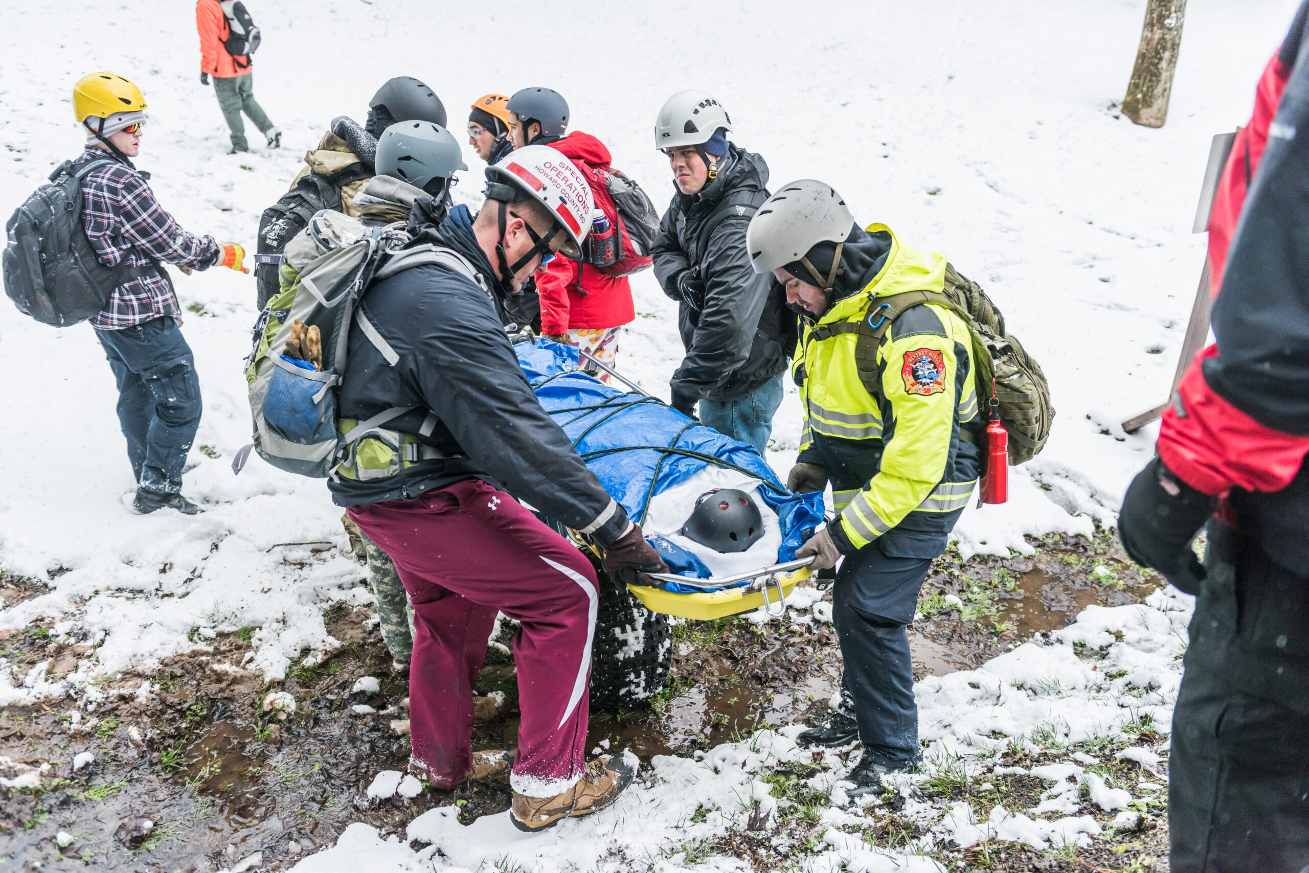 A group of people wearing winter gear and helmets carry a stretcher with a fake body. disaster health systems emergency