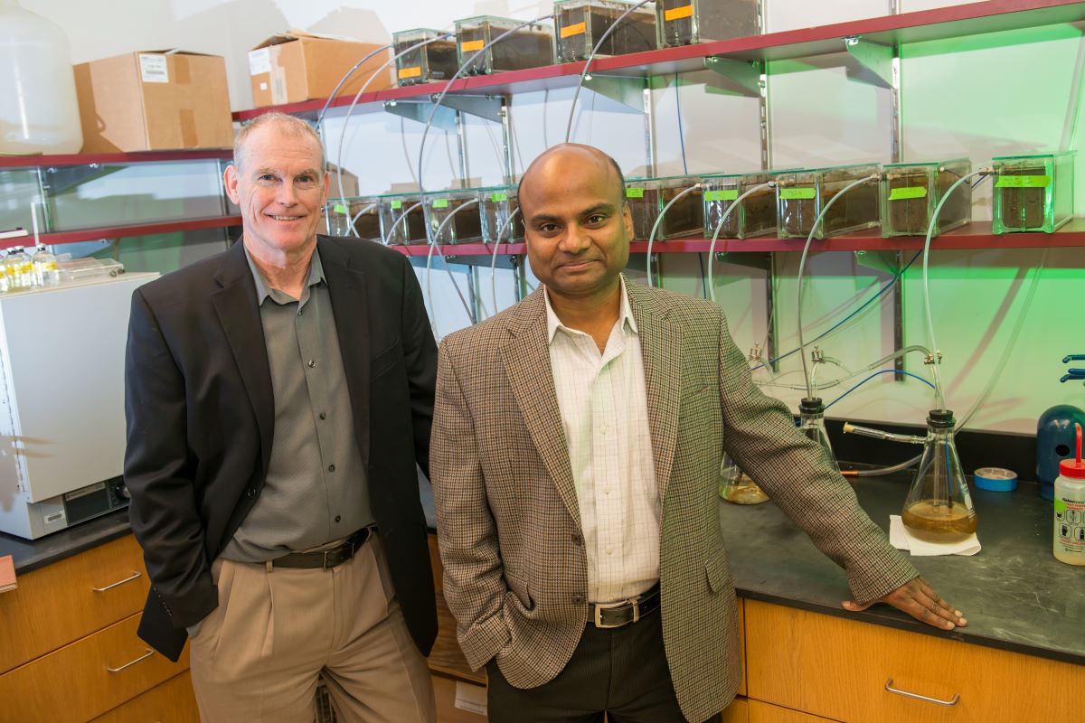 Two men wearing blazers, in front of lab bench with beakers and tubes, look at camera.