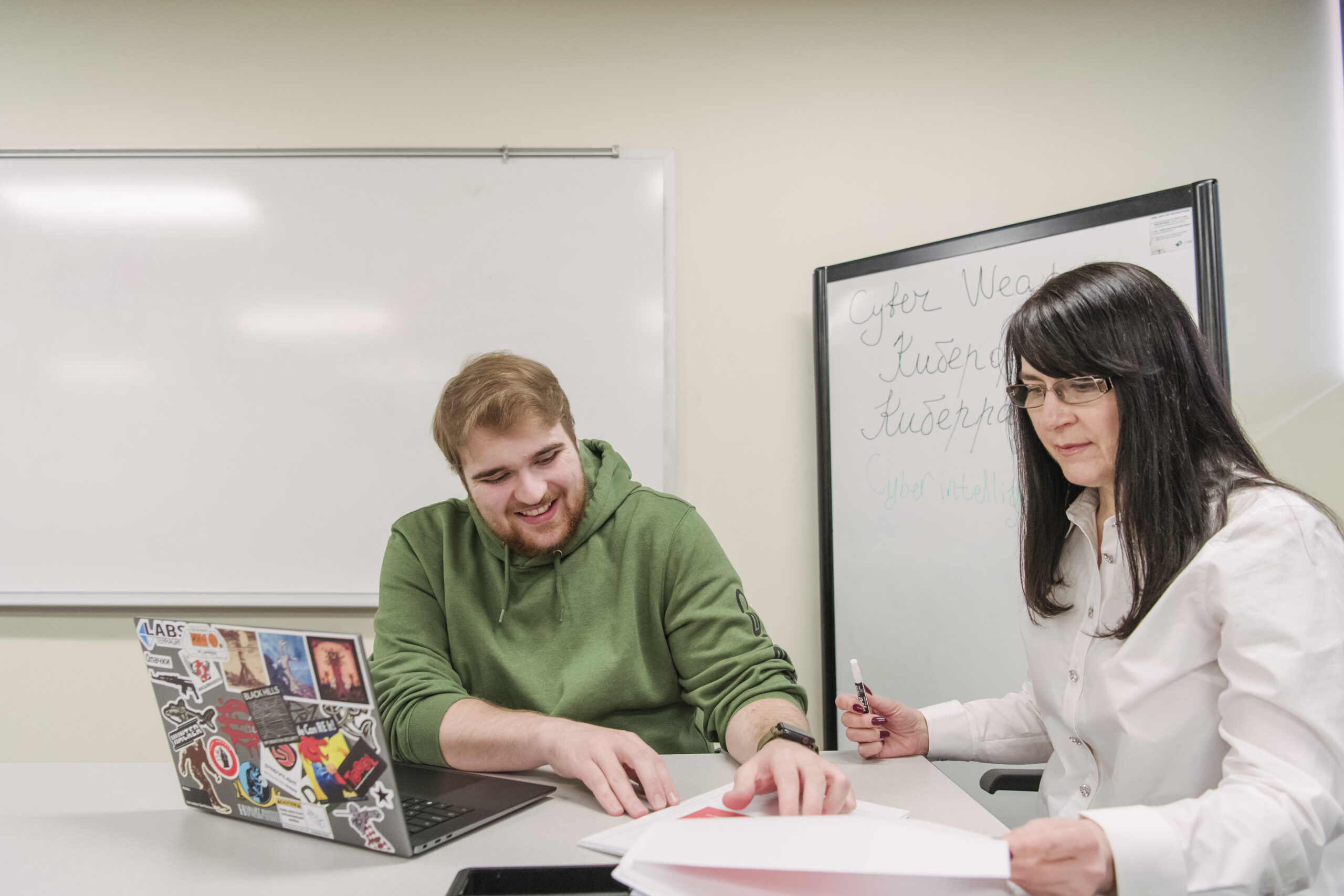Man and woman sitting at a desk in a classroom in front of a whiteboard with Russian words, focused on the papers in front of them.