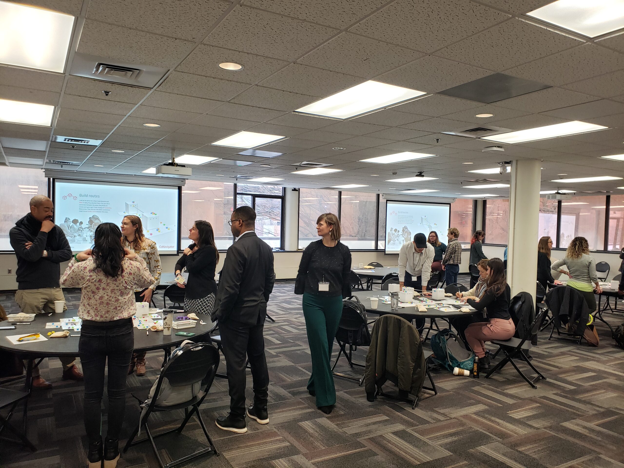 large multipurpose room with small groups of people clustered around round tables that are covered with notecards and small colorful tokens.