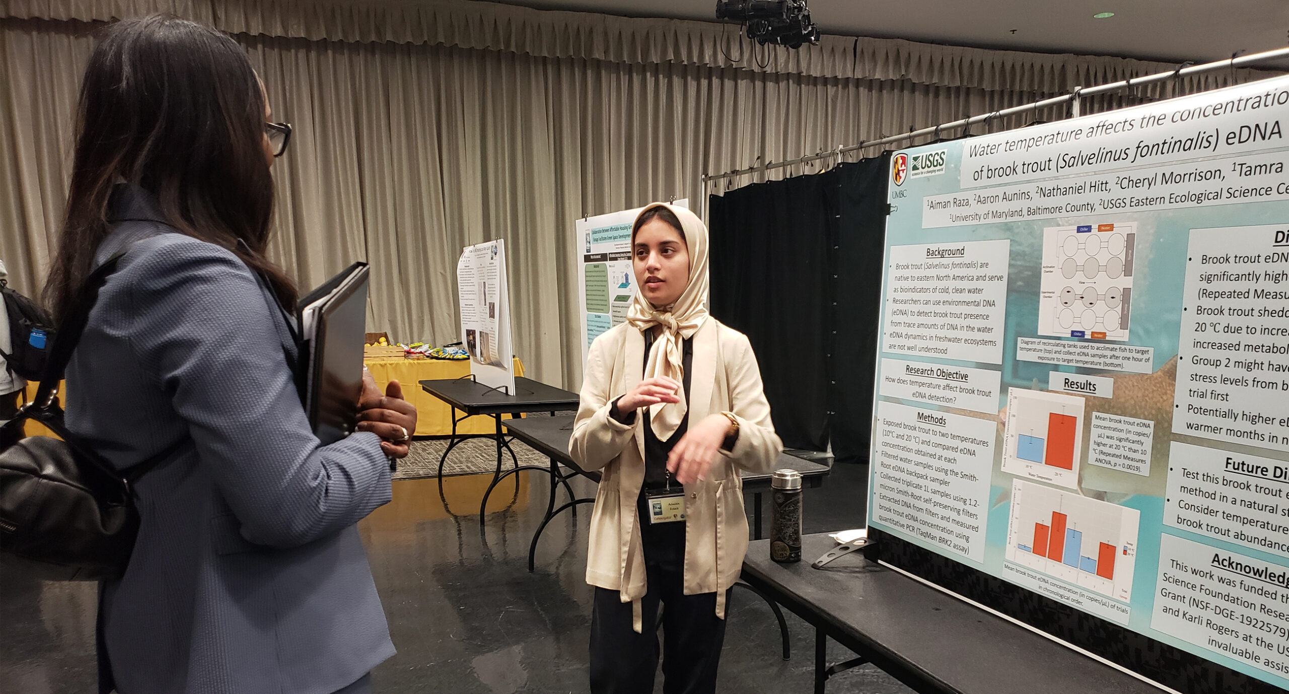 young woman stands near a research poster speaking to a standing woman in a high-ceilinged open hall