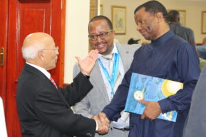 two men shake hands, one holding a large folder, while another looks on between them