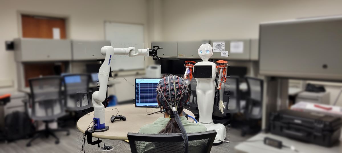 Woman wearing EEG cap sits with back toward camera. A computer screen, robotic arm and humanoid robot sit in front of her.
