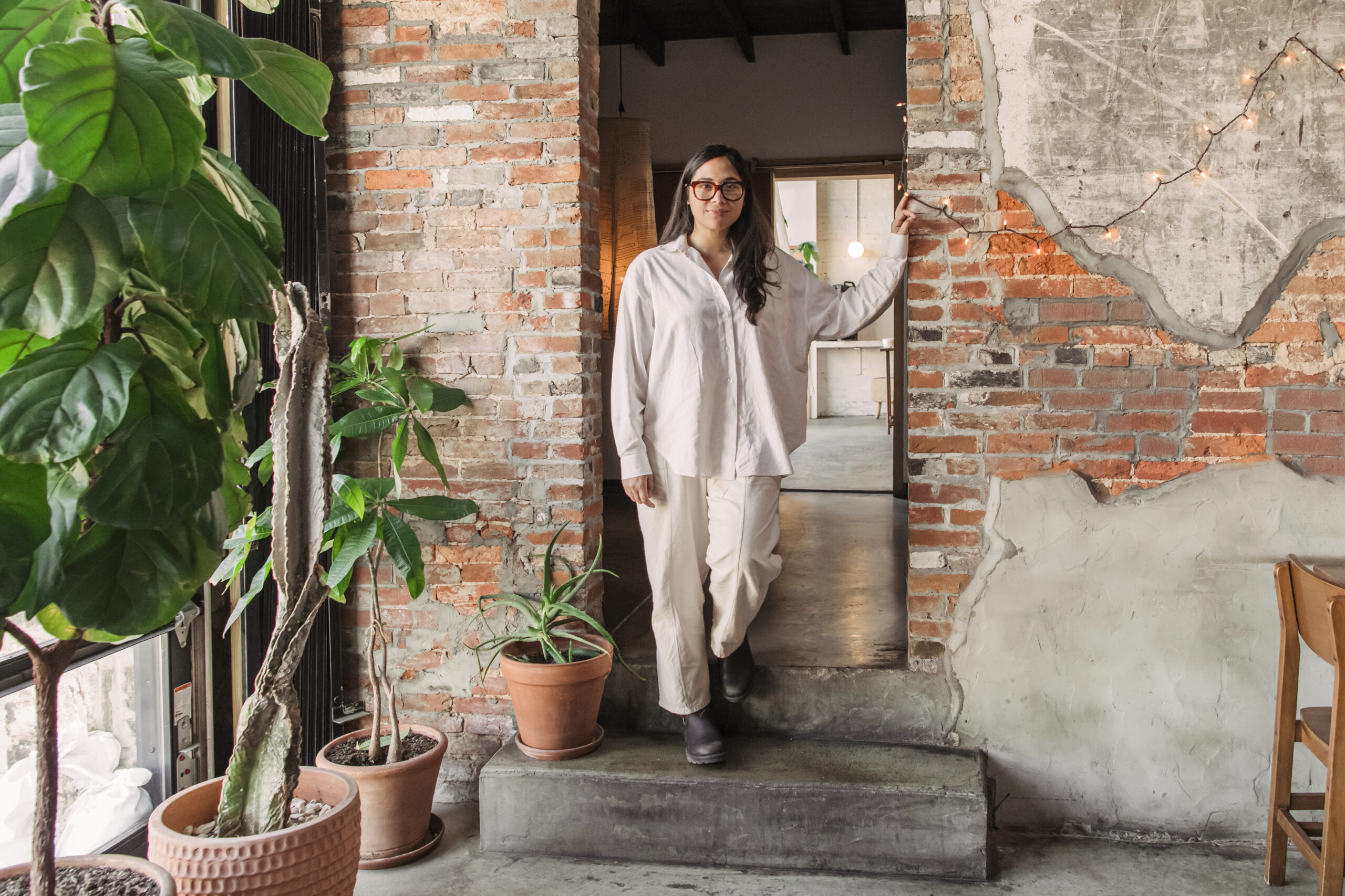 Harlan stands in a white and khaki outfit in a cutout of brick walls next to a green plant