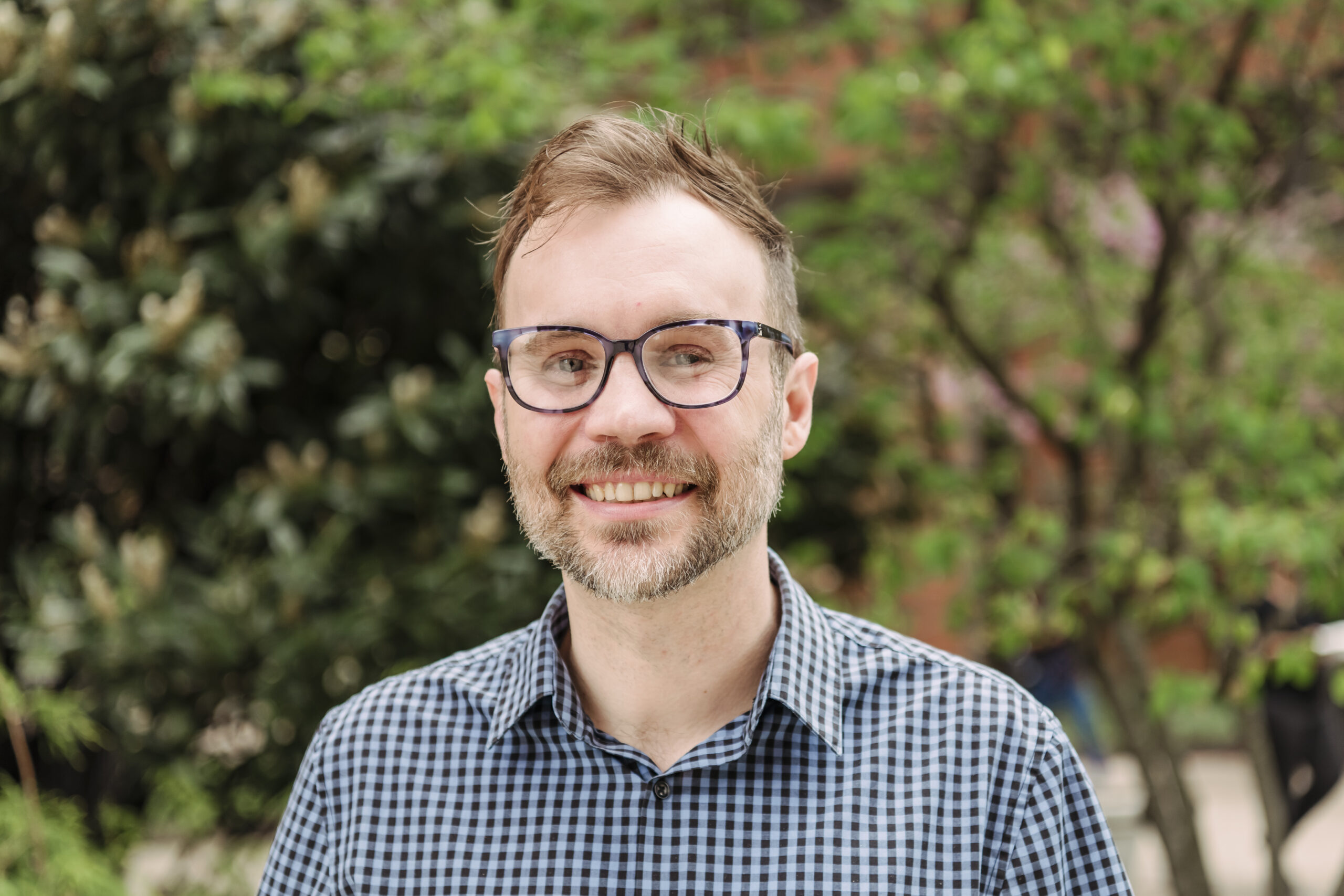 Man smiles at camera, greenery in background.