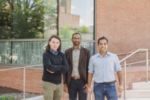 Three people pose for camera in front of brick building. Large window reflects the trees.