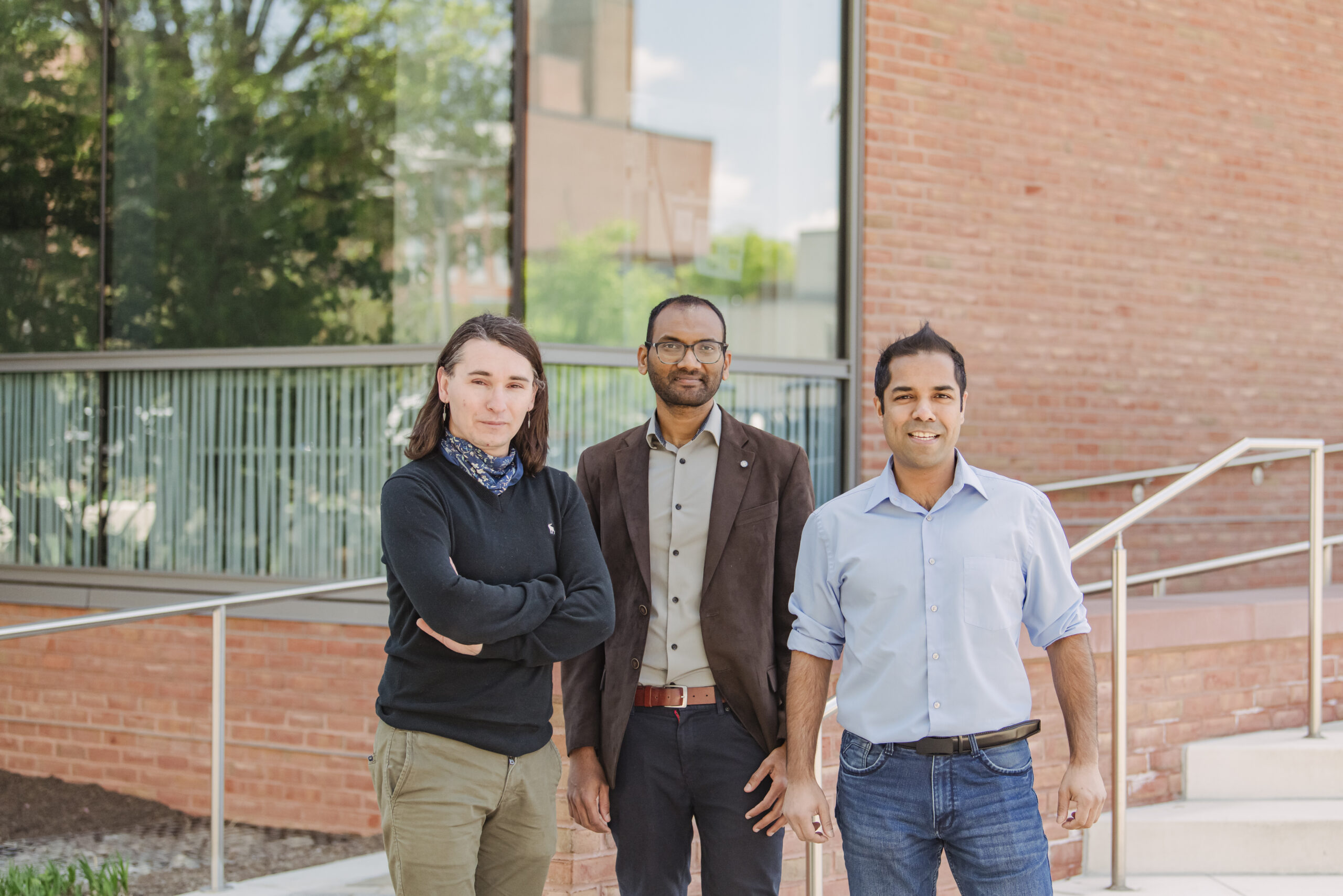 Three people pose for camera in front of brick building. Large window reflects the trees.