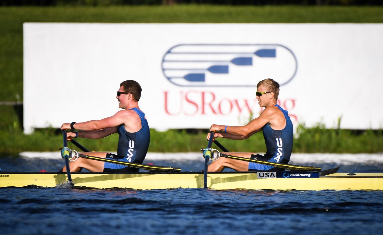 Two men in a rowing boat that is in water paddling oars in front of a backdrop that says USRowing