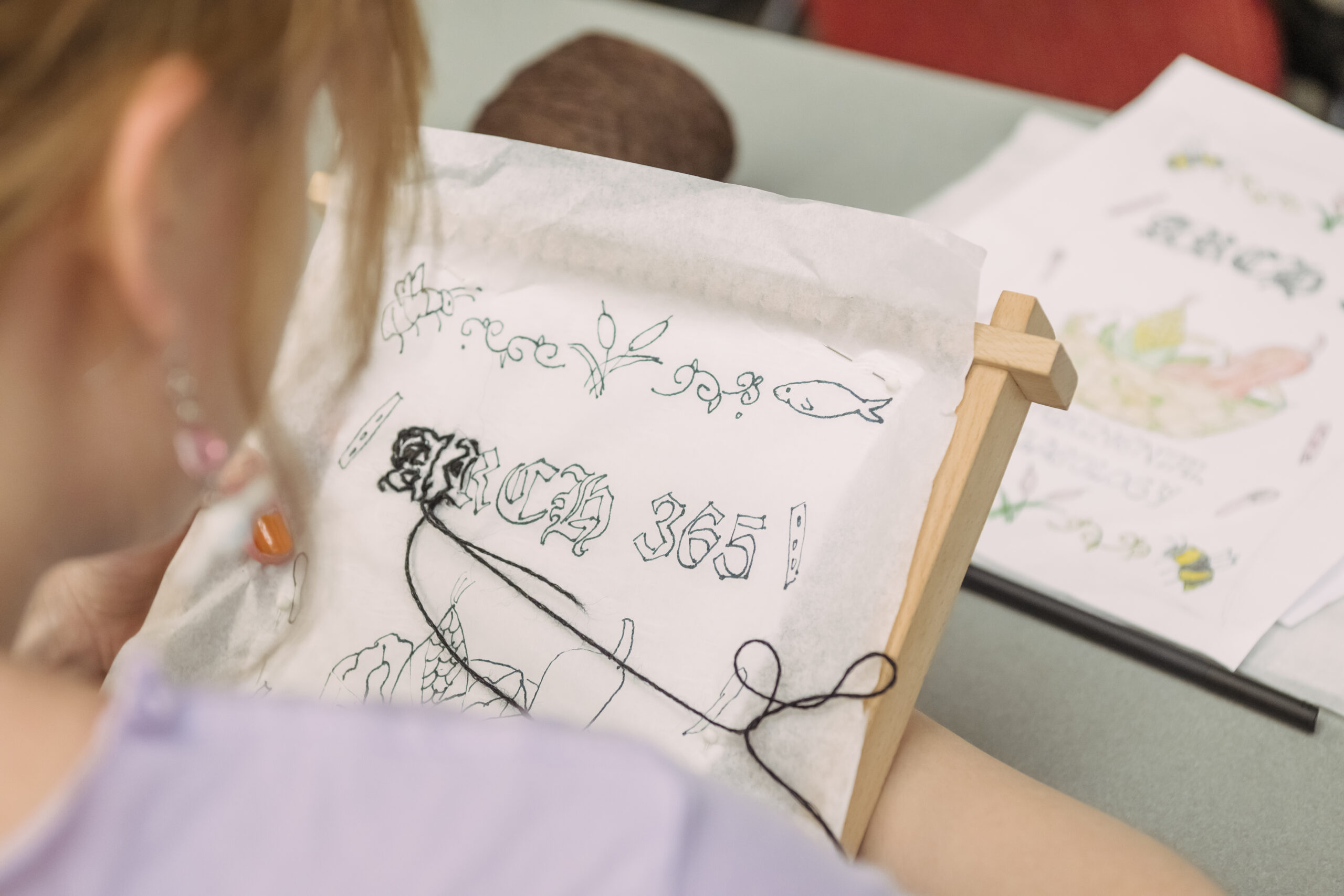 student actively stitching an embroidery project, which reads "ARCH 365" in a medieval font style