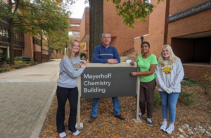 One professor and three students stand around a building sign that reads "Meyerhoff Chemistry Building" outside on a fall day, backed by a walkway and brick buildings
