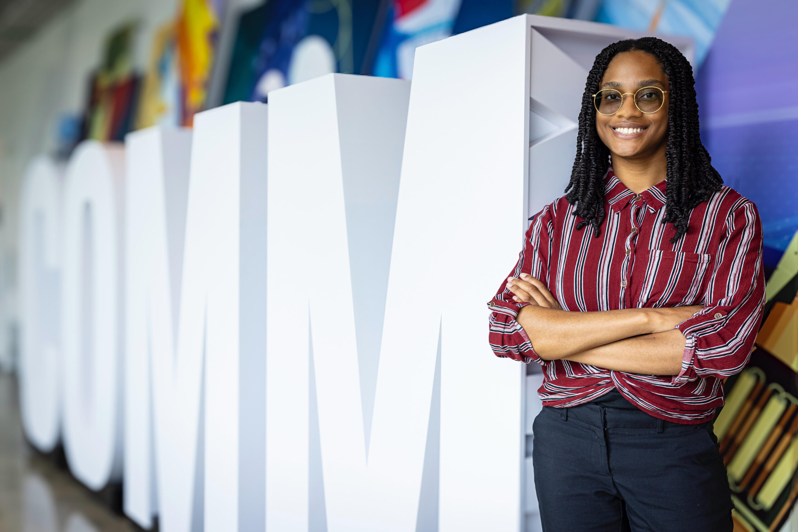 a young woman stands with glasses and her arms crossed in front of large cut out letters that spell COMM