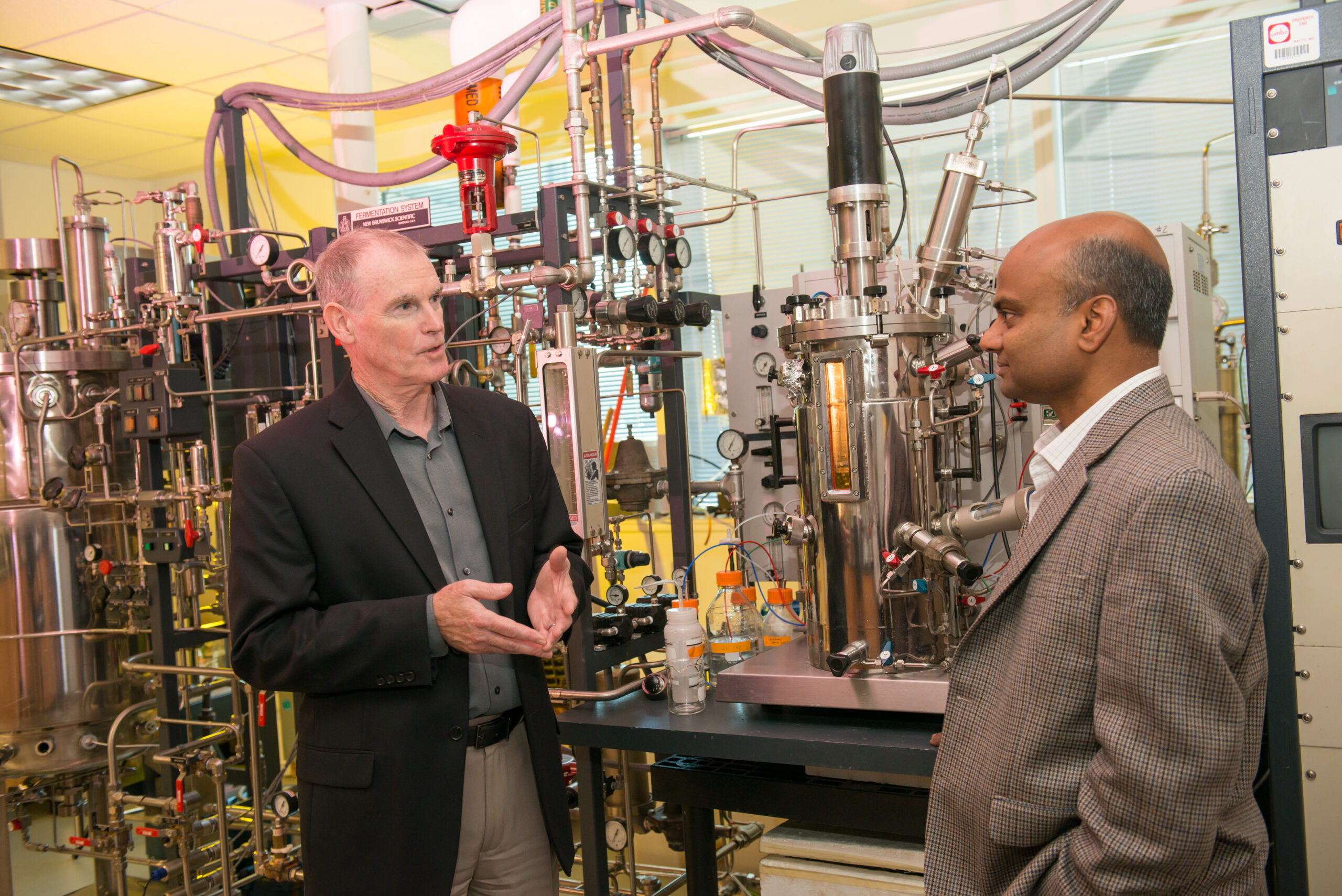 two men in conversation in front of a complicated set of tubes, flasks, and sensors