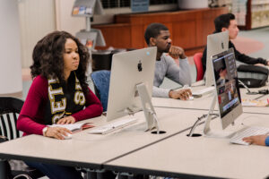 Students sit at table in front of computers. One wears a scarf that says "Retrievers"