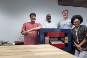 Four people pose in lab near structure with large metal components (magnet).