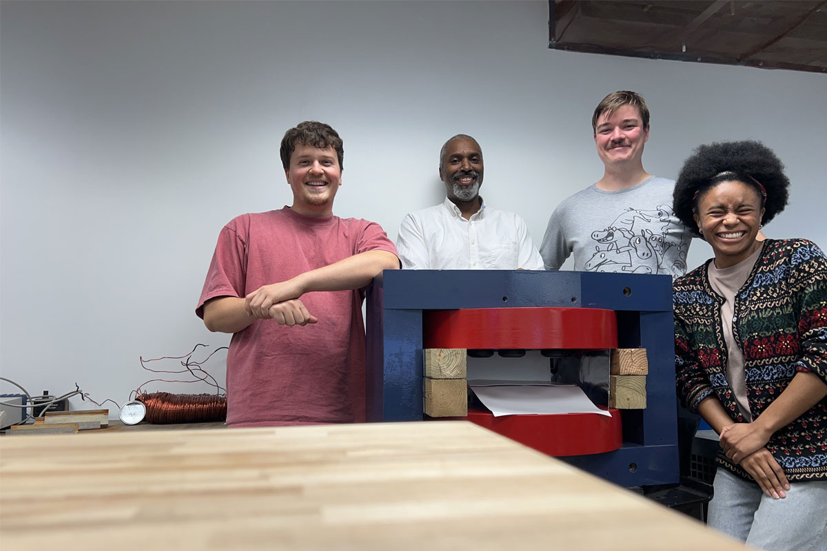 Four people pose in lab near structure with large metal components (magnet).