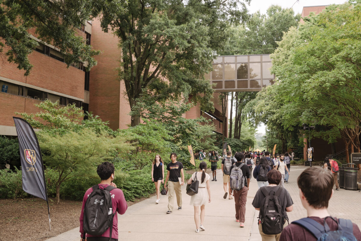 Students walk along pathway between brick academic buildings, trees on either side.