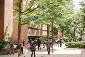 students walk on a pedestrian pathway between brick academic buildings, trees with spring leaves grow in adjacent beds