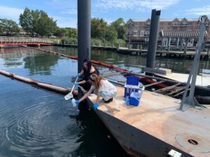 Two people, one an ICARE trainee, crouch on a pier near the water. One person pours water from one container into a bottle that the other person holds.