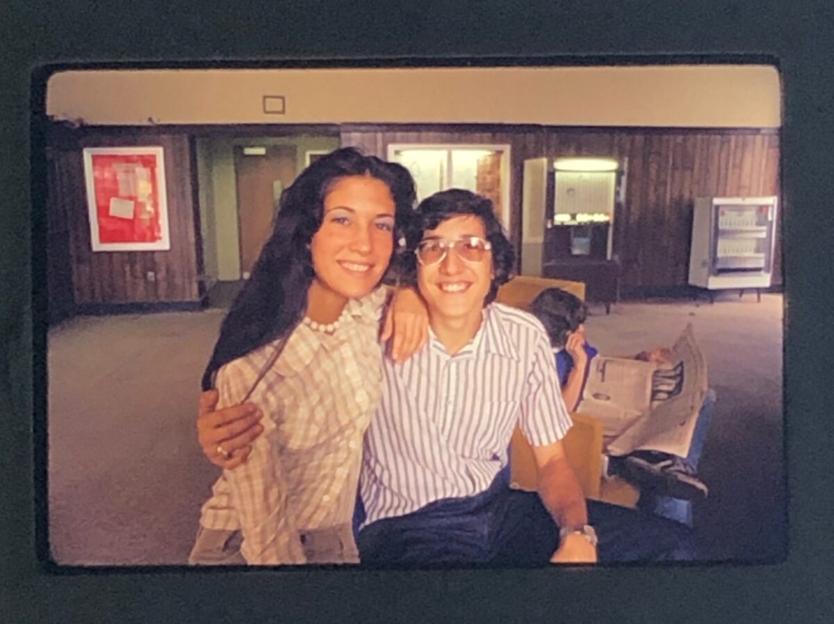 A photo from the 70s of a young man and woman smiling and sitting together in a room with wood-paneling and vending machines.