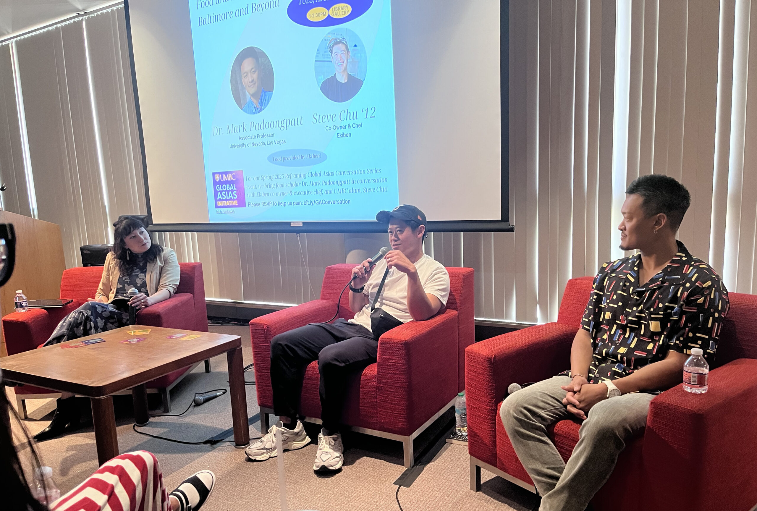 three people sit in red comfy chairs in front of an audience talking