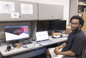 A man sits next to table with computers, electronic wiring and a robotic hand.