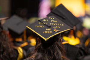 A decorated mortar board with the phrase "To all the plot twists that let me here" in gold glitter