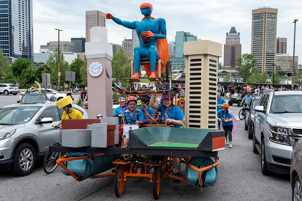Kinetic sculpture with teal man in wheelchair and cape, pedaled by team wearing blue shirts
