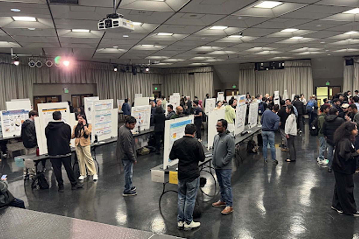People mingle in a large room around posters mounted on tables.