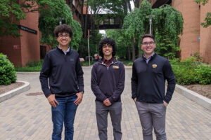 Three students wearing "UMBC" shirts stand outside, with academic buildings in background.