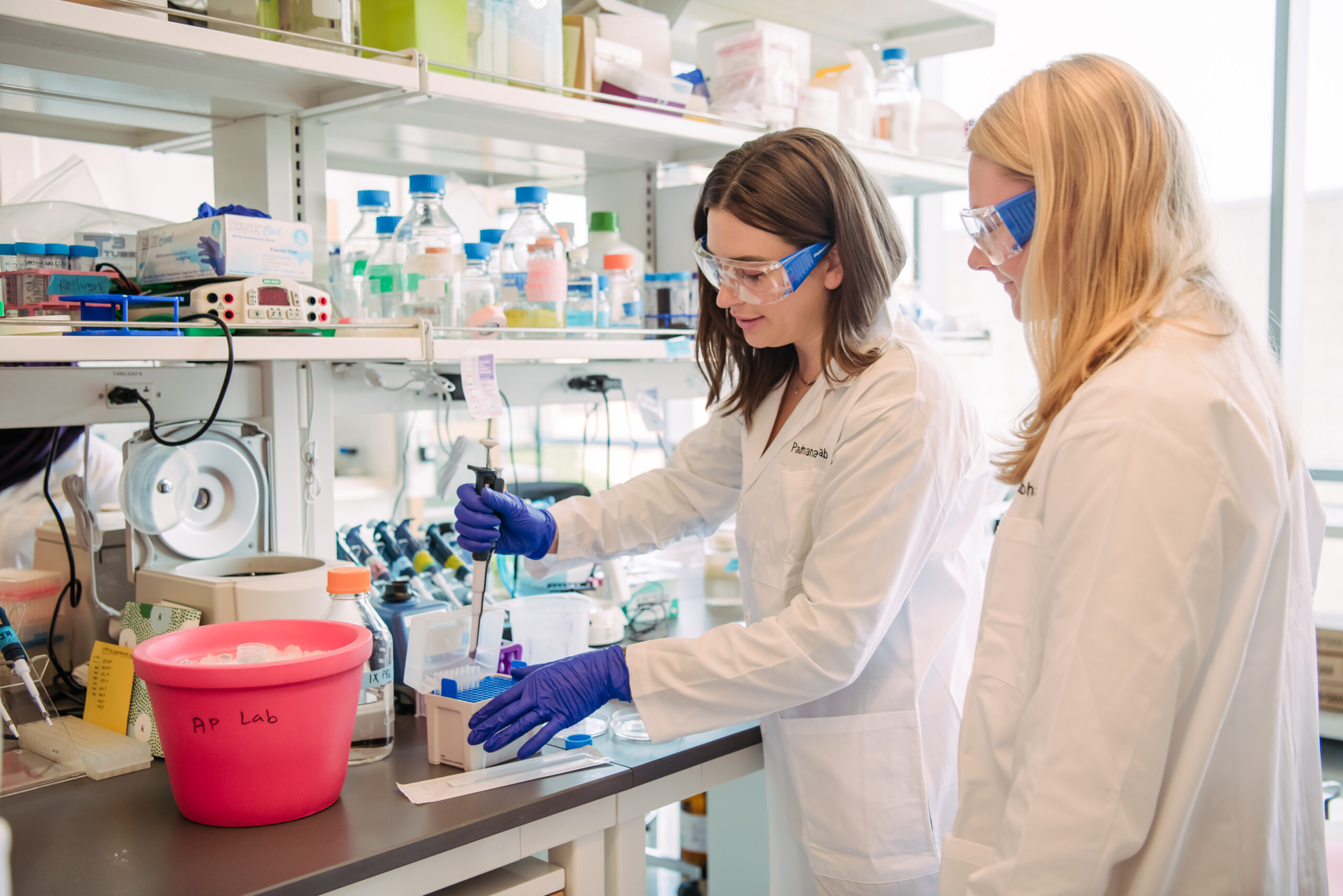two women in lab coats, gloves, and safety glasses in a brightly lit laboratory; one is pipetting while the other observes