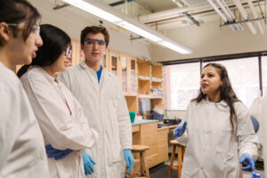 woman addresses three students, all wearing lab coats and gloves, in a brightly lit laboratory