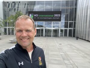 A man in a UMBC shirt takes a selfie in front of a conference center with a sign reading "HCI International 2023, 23-28 July, Copenhagen, Denmark" over the door