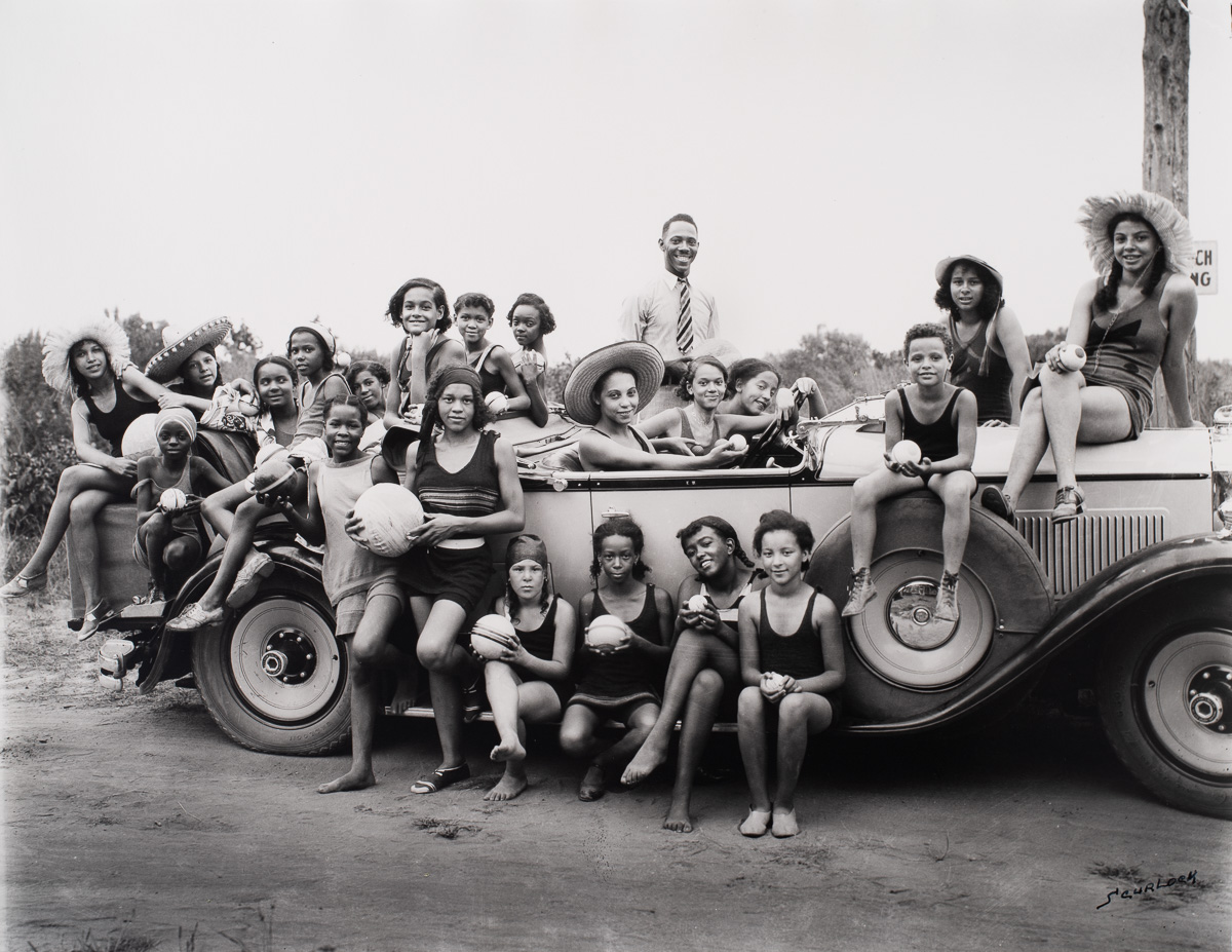 In a black and white photograph, a group of 22 Black Americans pose in and on a fancy early 1920s touring car.