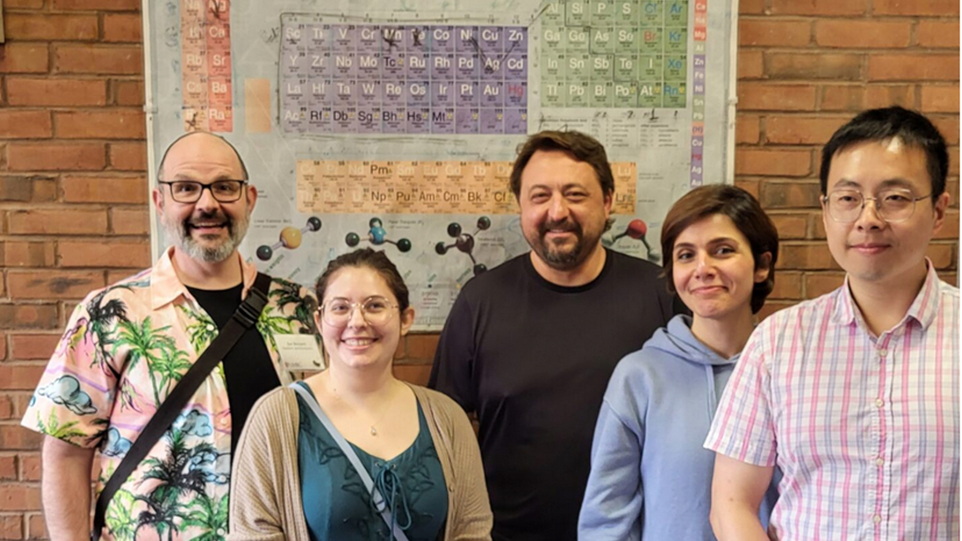 group photo of five people in front of a brick wall with a poster of the periodic table of the elements on it