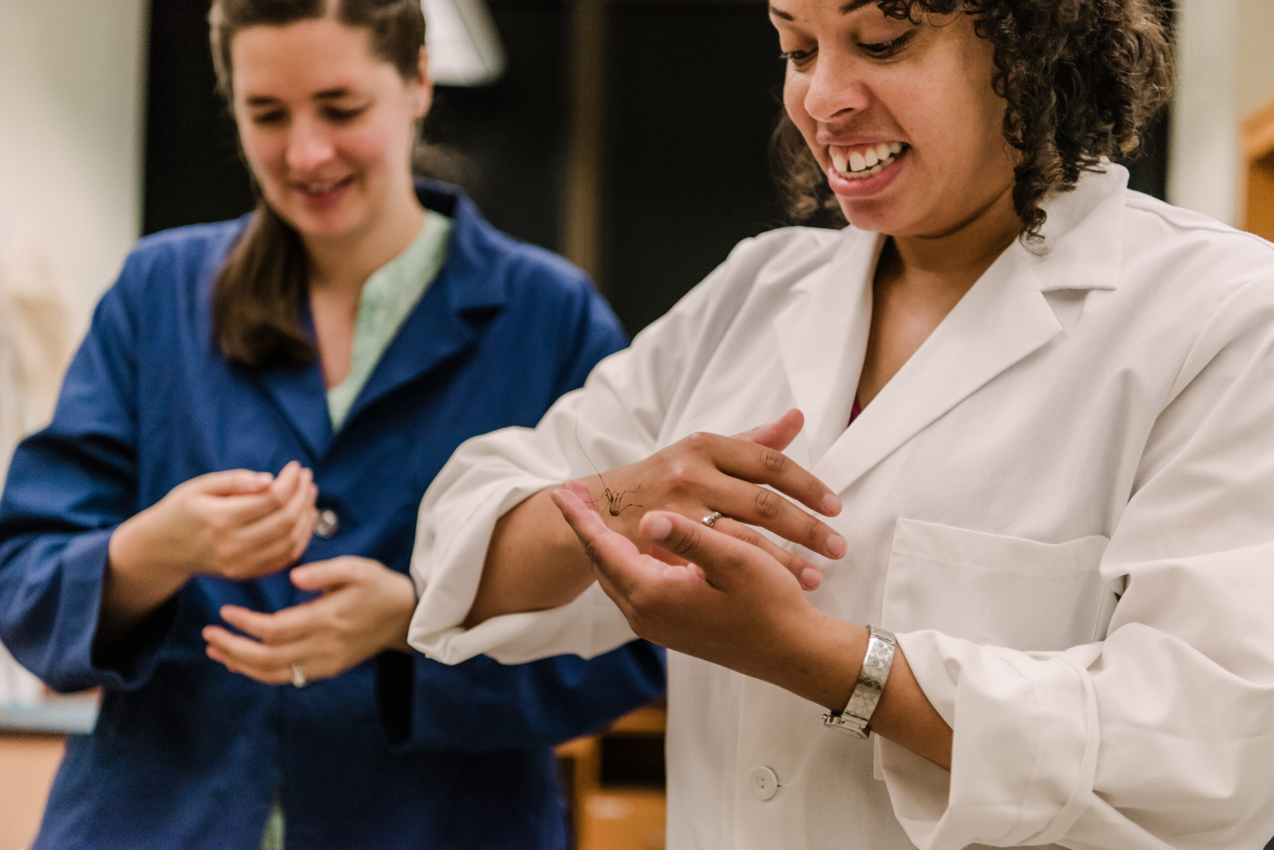 two women in lab coats; one smiling with a daddy longlegs crawling on her hands