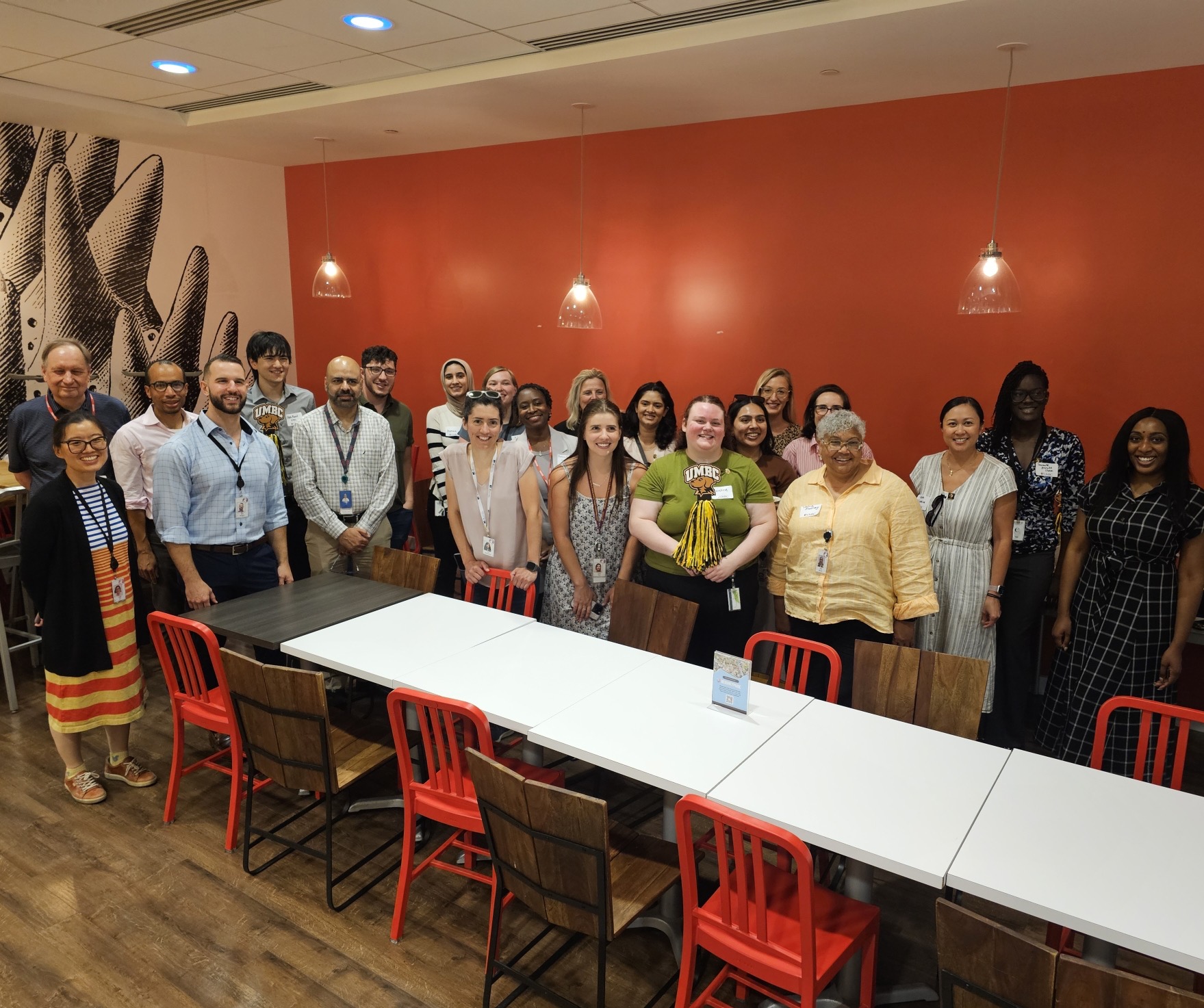 large group photo in front of a red wall, behind a long rectangular table with red chairs
