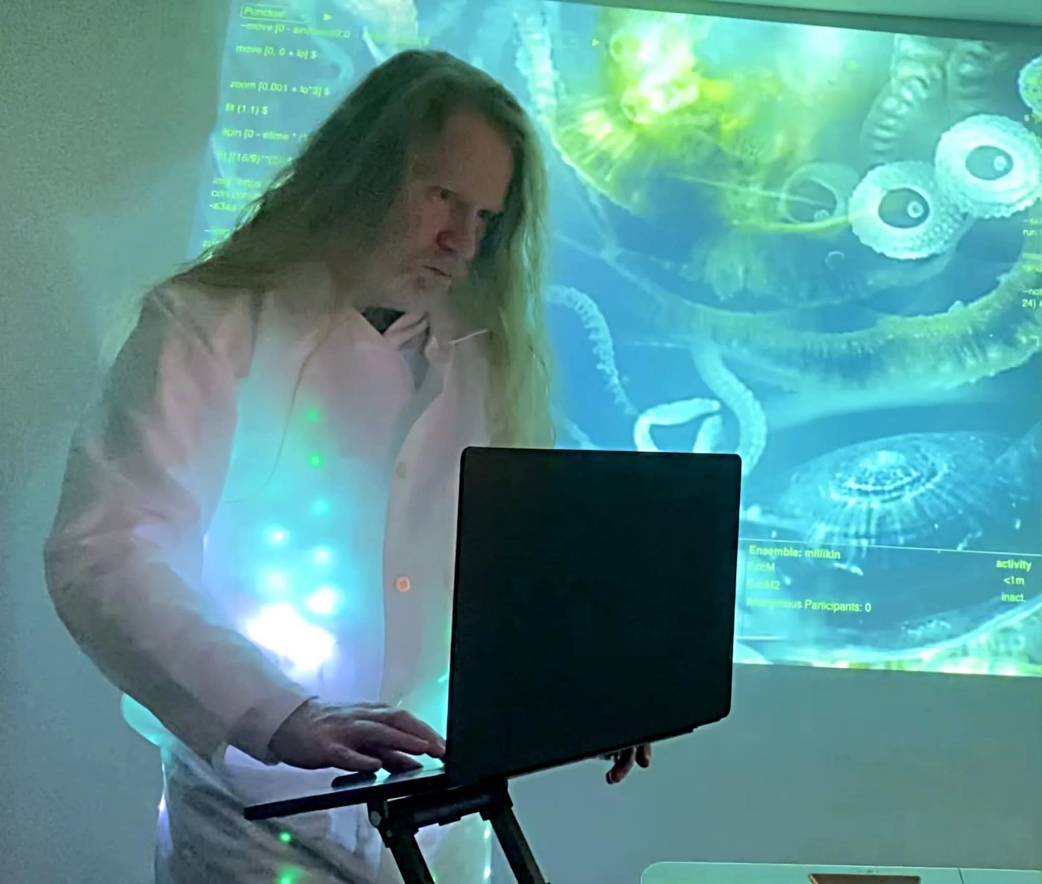 Artist Eric Millikin stands with a laptop in front of a screen filled with abstract blue and green shapes.