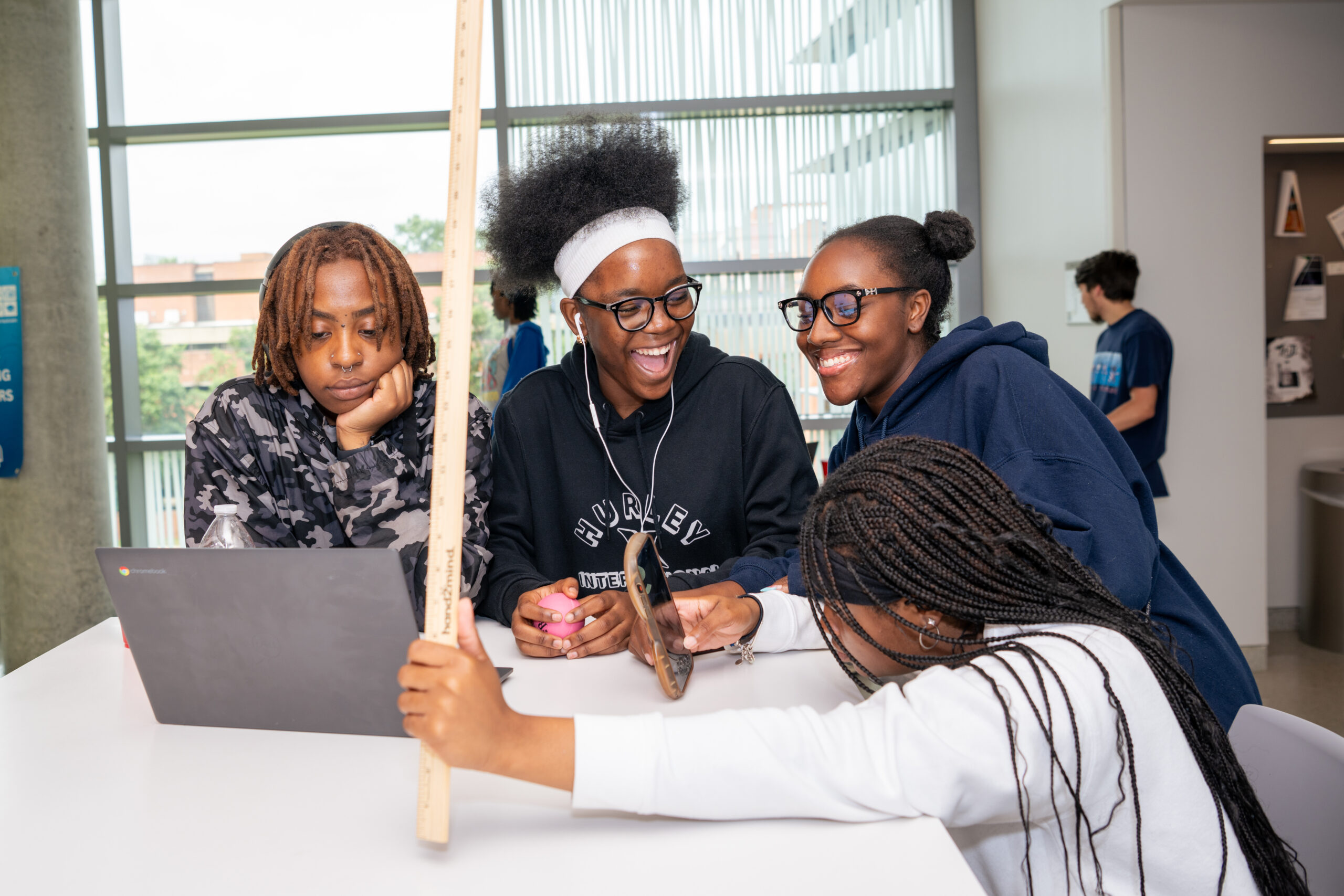 four high school students smiling and laughing around a white table; a laptop sits on the table and one student is holding up a meterstick