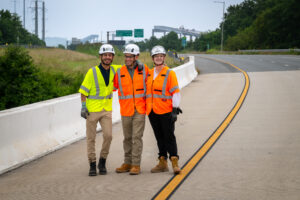 Three students wearing hard hats and reflective vests stand on a road. The missing span of the Key Bridge is evident in the background.