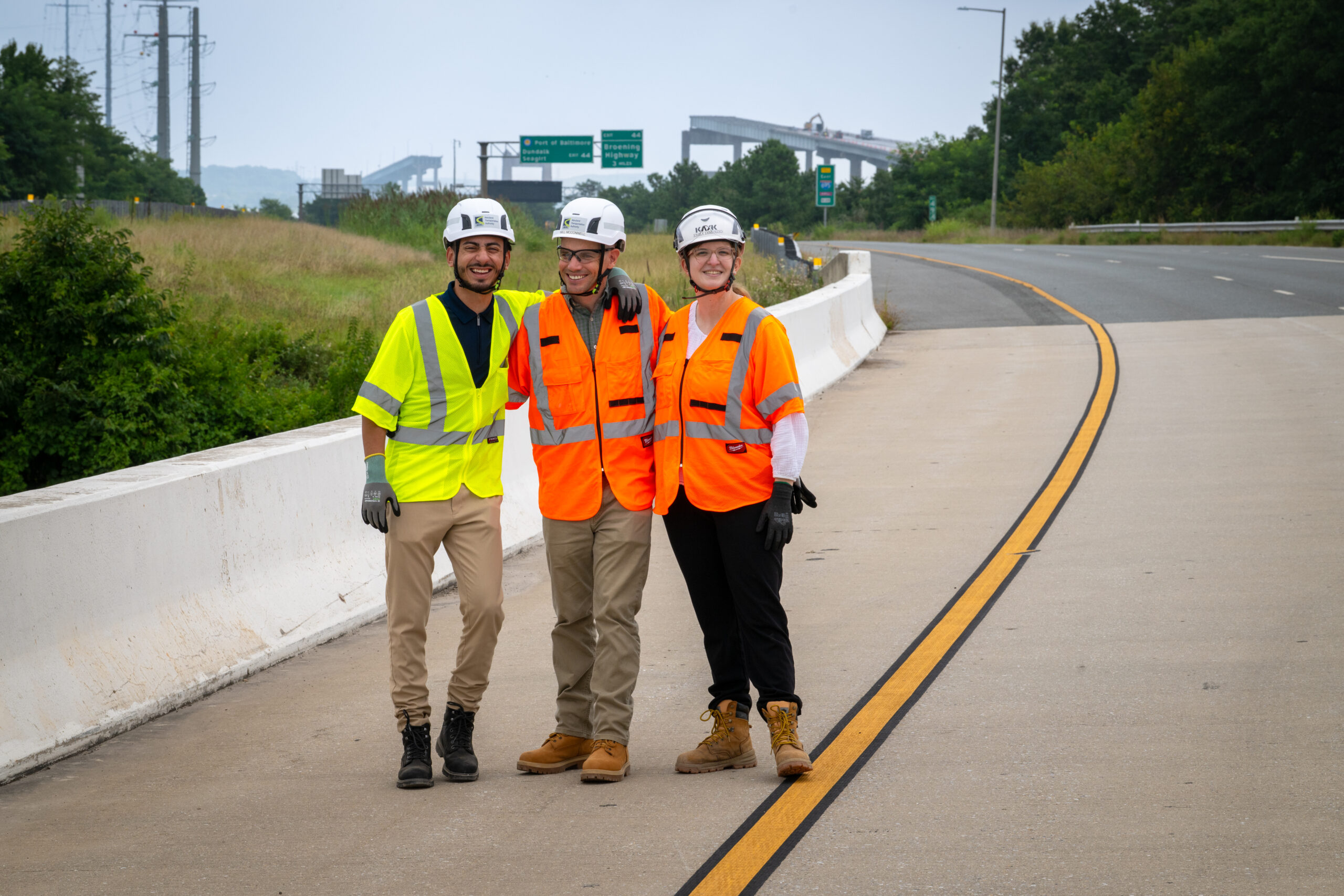 Three students wearing hard hats and reflective vests stand on a road. The missing span of the Key Bridge is evident in the background.