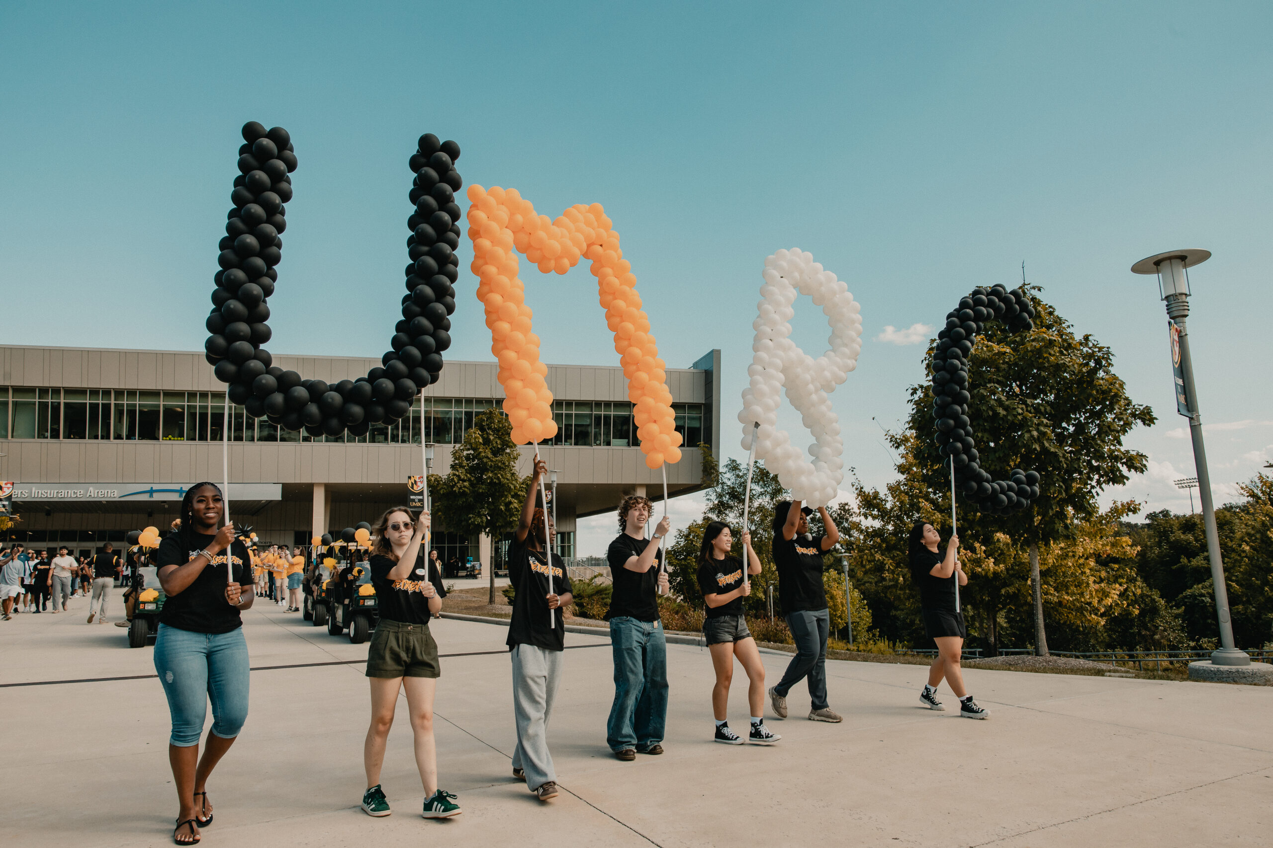 black, gold, and white balloons spell out "UMBC," with a student carrying each letter as they march in front of the CEI Arena