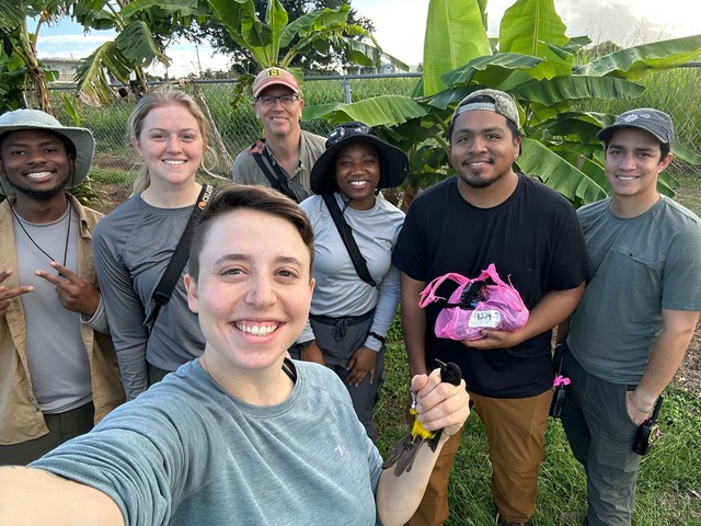 group photo of seven people, backed by tropical forest, one holding a songbird
