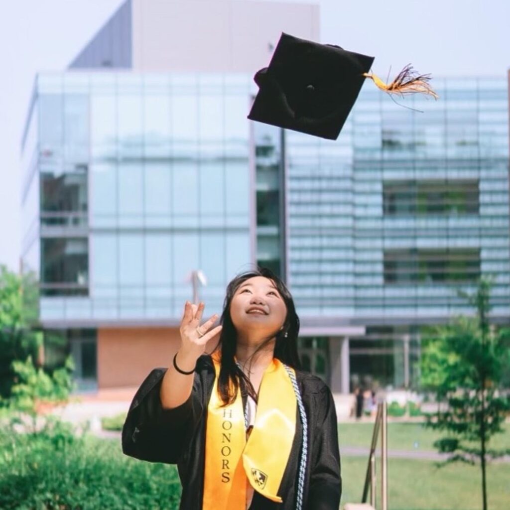 Ivy Nguyen, a college student wearing graduation robes and honors gold sash tosses her mortar board in the air