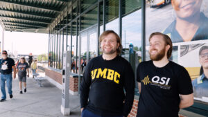 two people standing on a light rail platform; one's t-shirt reads "UMBC" the other "QSI"