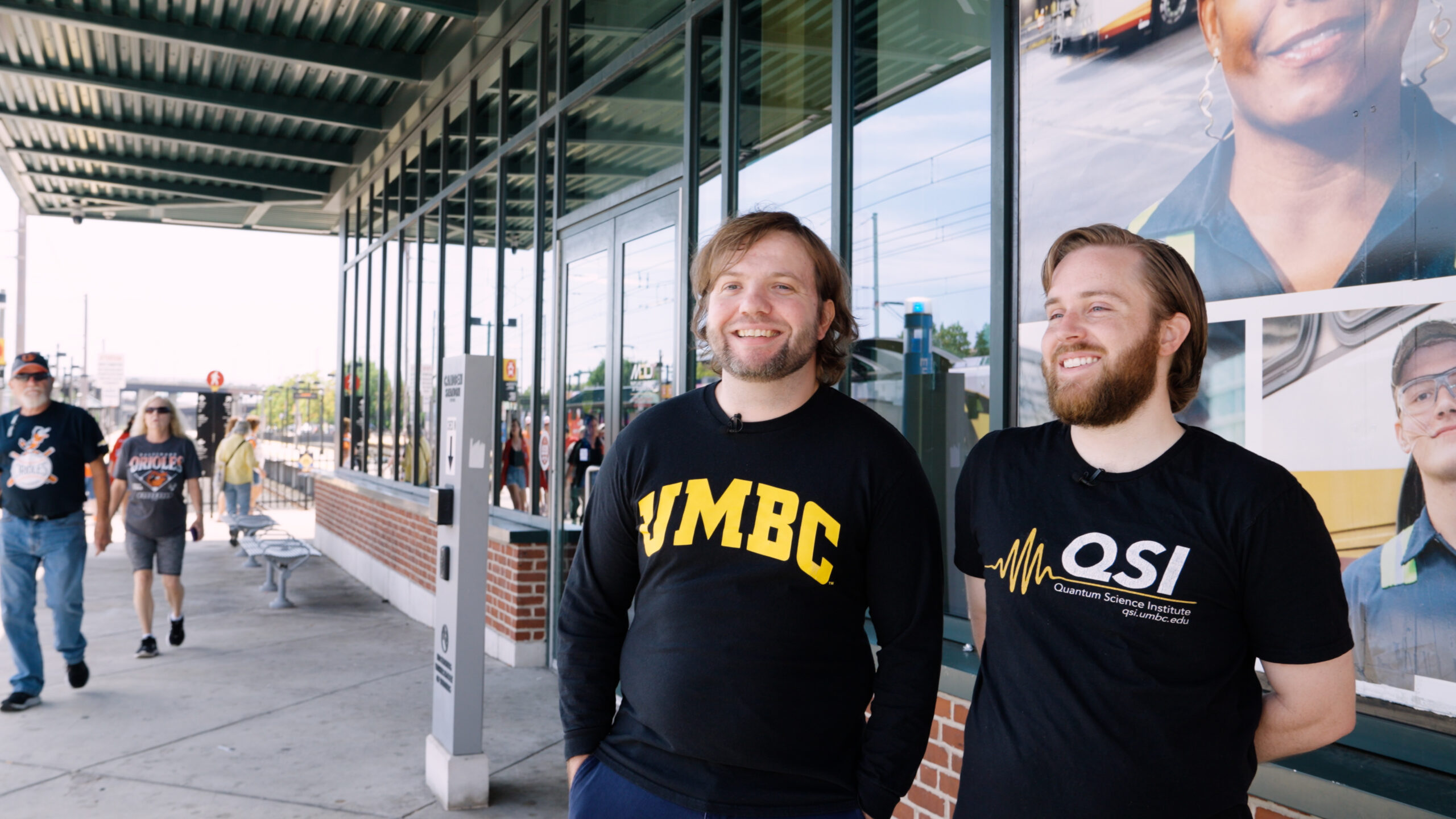 two people standing on a light rail platform; one's t-shirt reads "UMBC" the other "QSI"