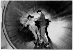 Black and white photo of a man and woman standing in a tunnel. The man is helping the woman stand, they are both smiling.